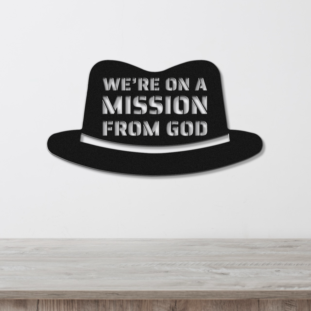 A metal wall sign with a fedora design and the text 'WE'RE ON A MISSION FROM GOD' in bold white stencil lettering against a black background.